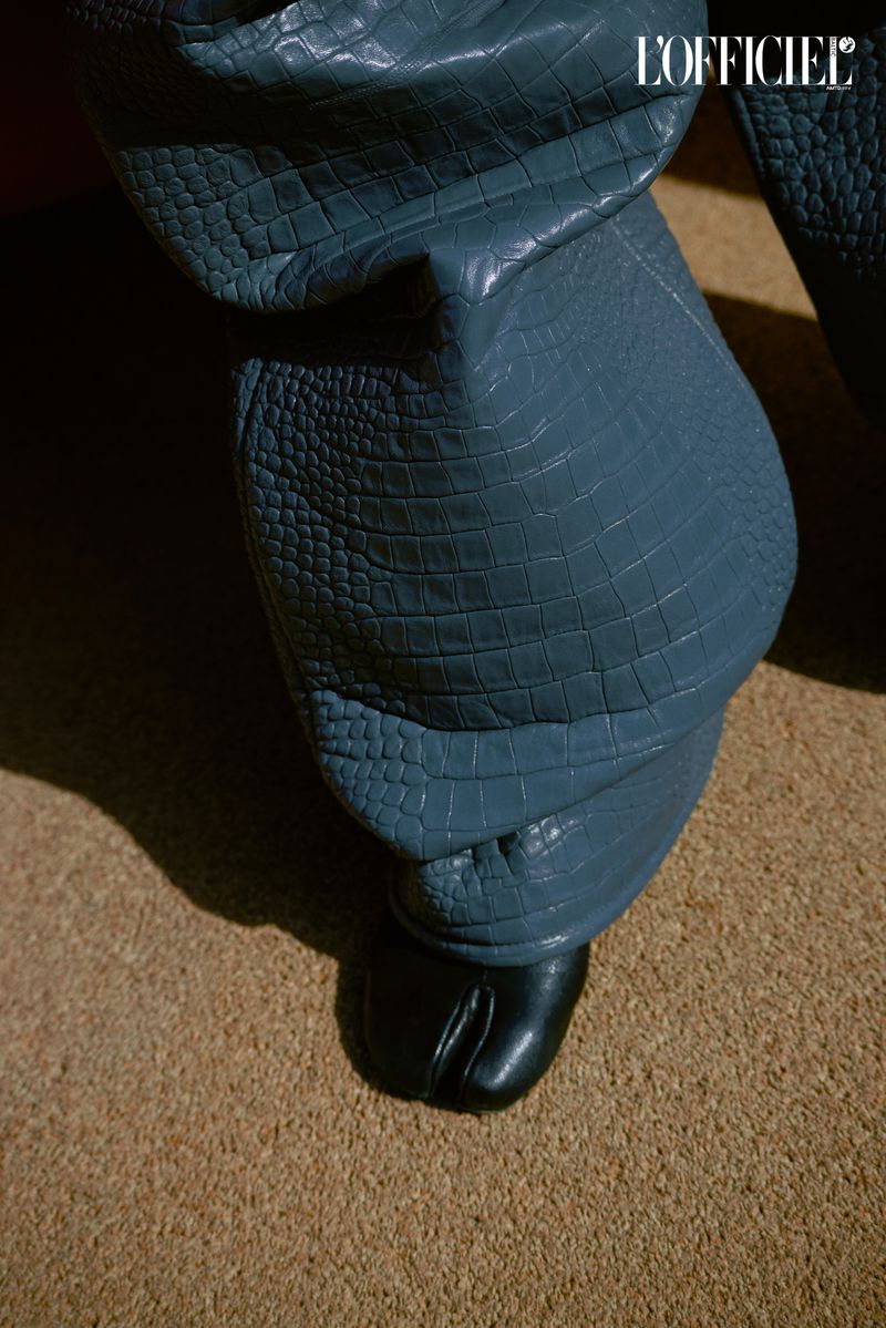 Close-up of blue faux alligator pants and black split-toe shoes on a beige carpet, featuring a logo in the upper corner.