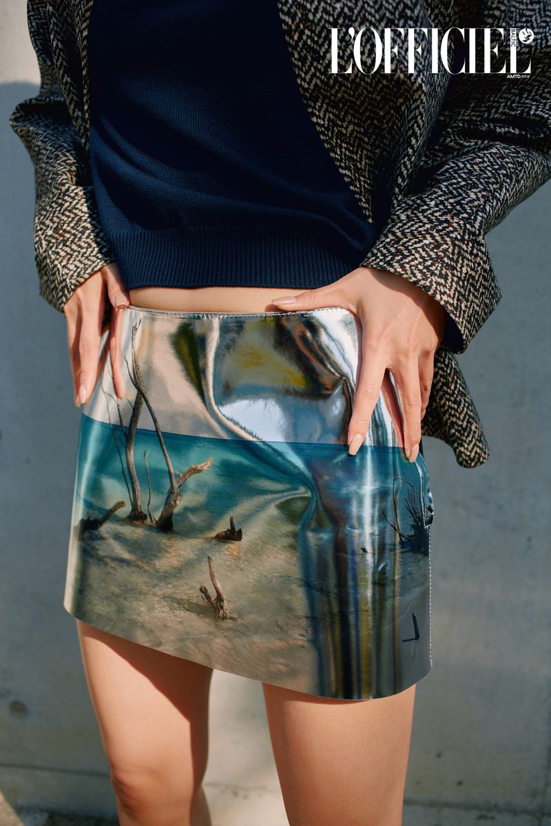 Fashion shot of a woman wearing a patterned skirt and blazer, standing indoors with a magazine logo visible in the upper corner.