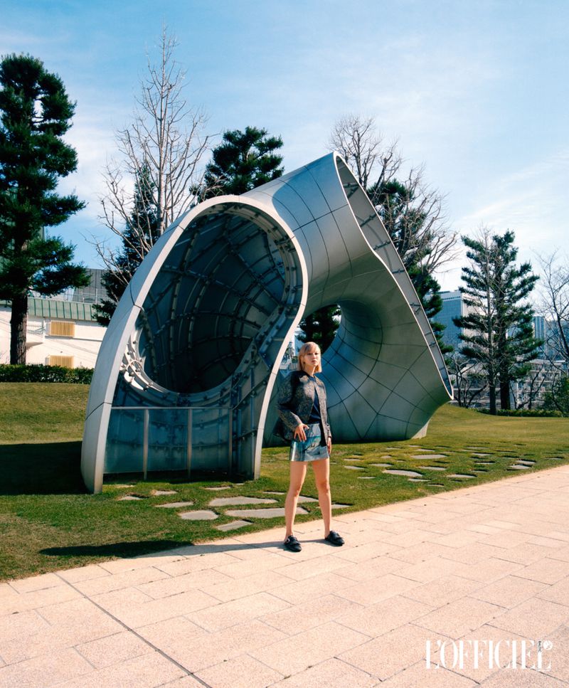 Young adult woman posing in an outdoor park in front of a metal sculpture, wearing a black jacket and denim shorts on a sunny day.