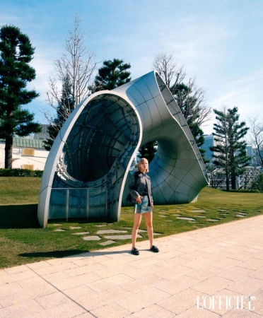Young adult woman posing in an outdoor park in front of a metal sculpture, wearing a black jacket and denim shorts on a sunny day.