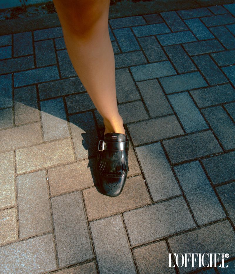 Fashion editorial shot: A woman's leg and black leather loafer with a silver buckle on a patterned brick surface.