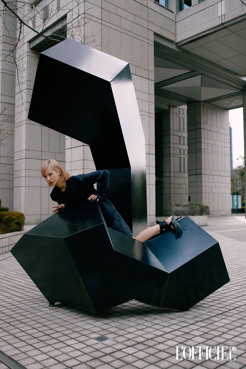 Fashion shot of a blonde woman in a dark outfit leaning over a large geometric sculpture on a paved surface with a building in the background.