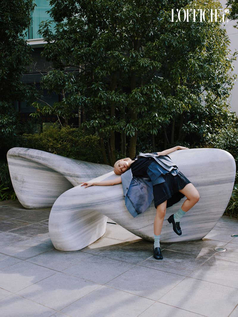 Fashion editorial shot of a woman posing on a marble sculpture in an outdoor setting with trees and modern architecture in the background.