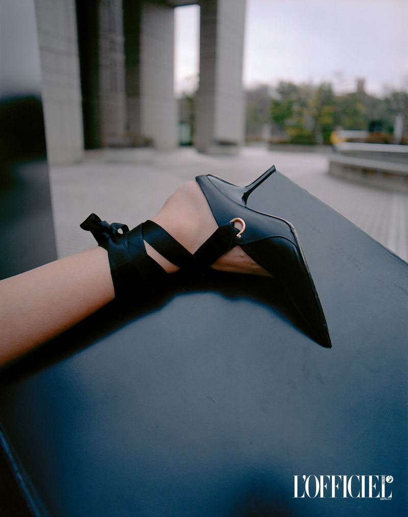 Close-up shot of a woman's ankle and foot wearing a black, pointed stiletto with ribbon ankle ties, against a gray architectural backdrop.