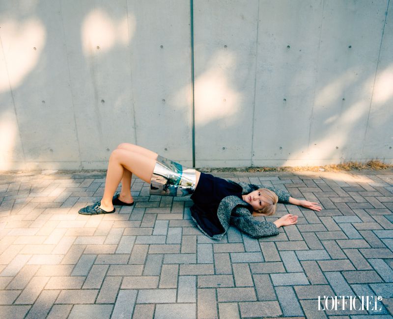 A fair-skinned woman with blonde hair in an arched back pose wearing shiny shorts and a sweater lies on a stone-paved surface outdoors.