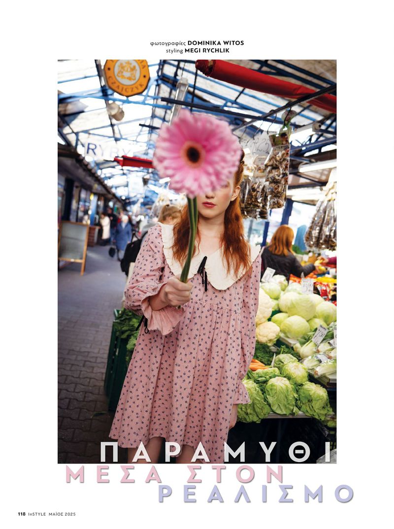 A red-haired adult female model stands in an open market, holding a large pink gerbera daisy in front of her face, wearing a polka dot dress.