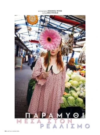 A red-haired adult female model stands in an open market, holding a large pink gerbera daisy in front of her face, wearing a polka dot dress.