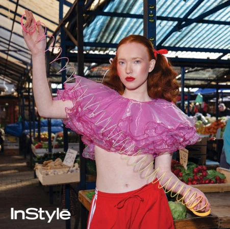A woman with red hair poses in a market, wearing a pink top with white zigzags and red shorts, accessorized with coiled bracelets.