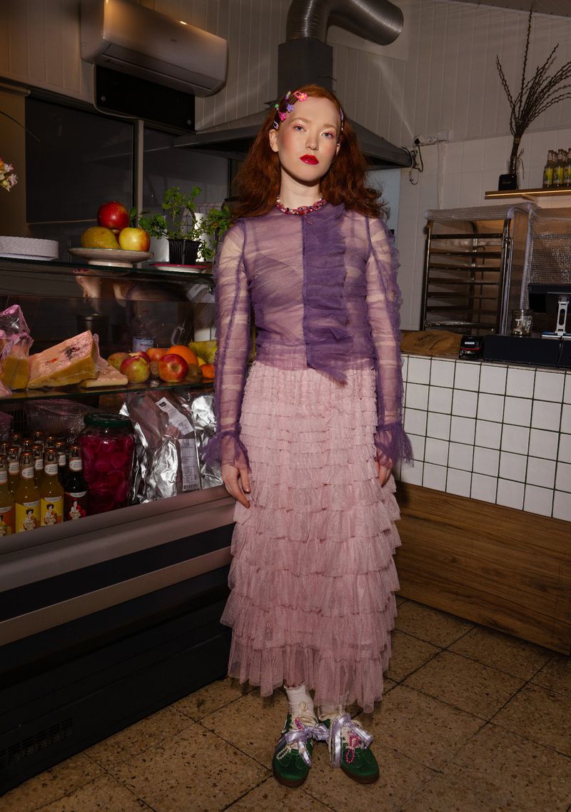 Full-length portrait of a red-haired woman in a ruffled purple top and tiered pink tulle skirt, standing in a commercial kitchen.