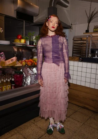 Full-length portrait of a red-haired woman in a ruffled purple top and tiered pink tulle skirt, standing in a commercial kitchen.