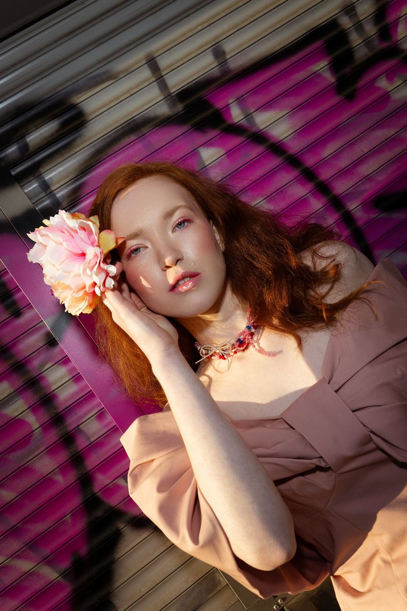 Adult female fashion model with red hair and flower accessory poses near graffiti wall in off shoulder top and jewelry necklace.