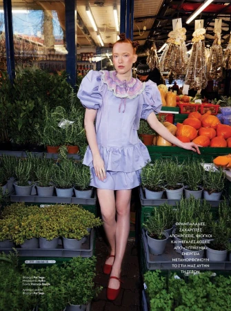 A young woman in a lavender dress stands in a market filled with greenery and produce, creating a vibrant and fashionable portrait.