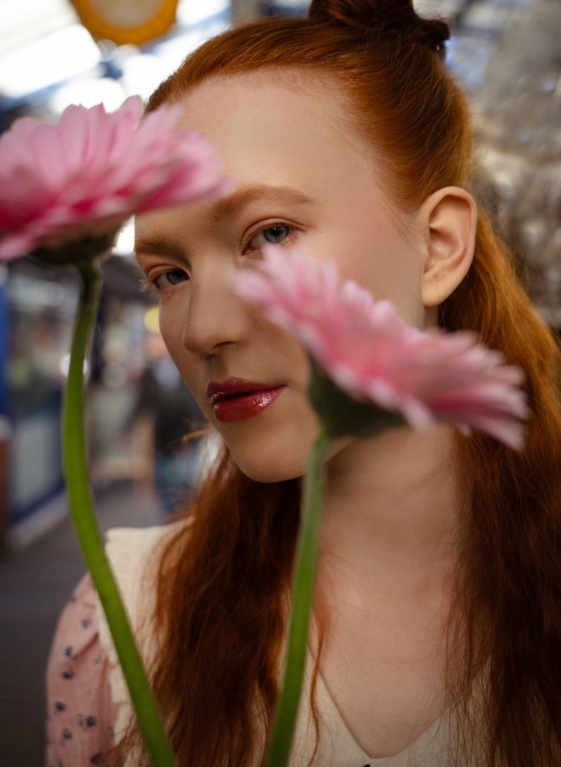 Close-up of a woman with red hair posing behind two pink gerbera daisy flowers in an indoor market, soft focus and diffused lighting.