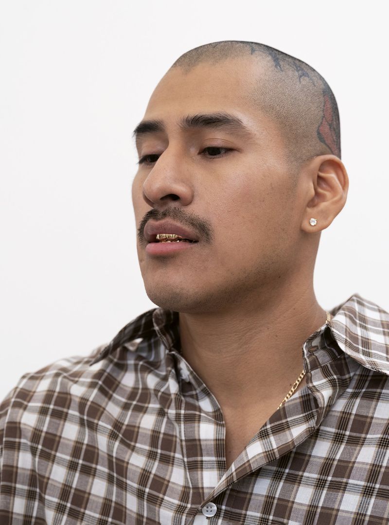 Portrait of an adult male wearing a brown and white plaid shirt. He has gold teeth and a gold chain. The background is white.