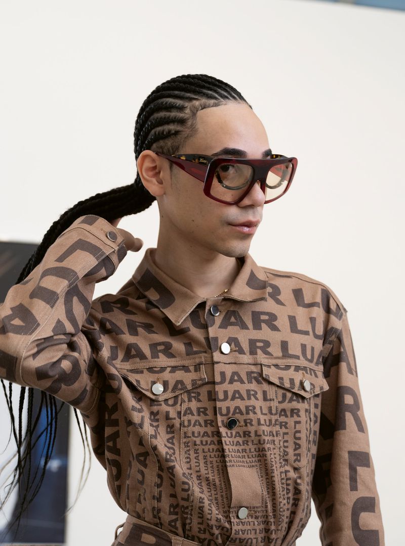 Stylish individual with braids and oversized glasses wearing a LUAR logo jacket poses in a bright indoor setting with a casual expression.