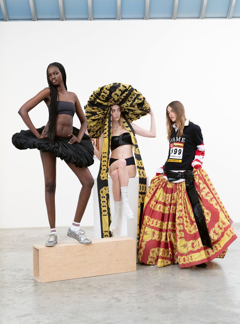 Three fashion models pose indoors against a white wall, wearing unique outfits, one wearing a large decorative hat.