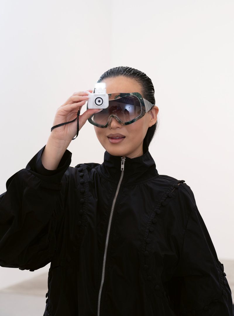 A woman in a black jacket poses indoors wearing oversized goggles and holding a white device over her eye in a brightly lit shot.