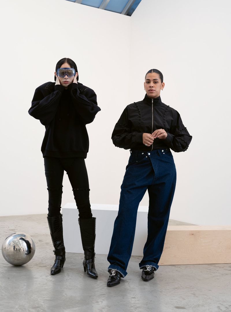 Two women stand in a minimalist white studio wearing avant-garde black outfits and blue denim. A silver sphere sits to the left.