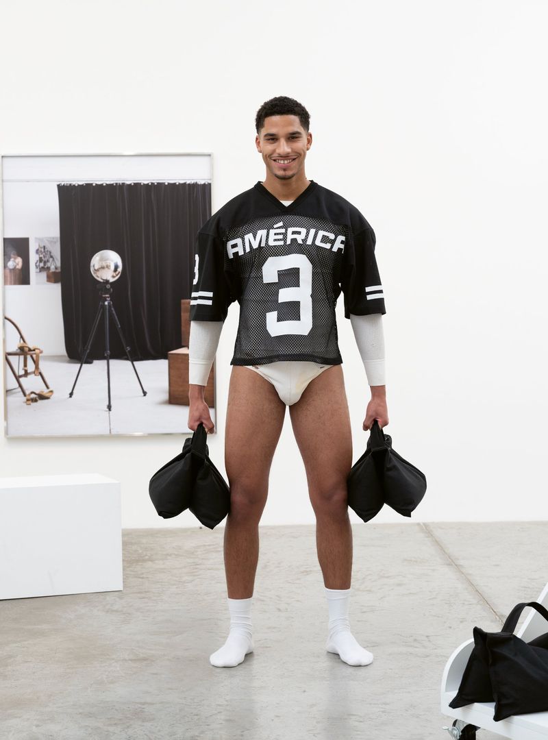 Athletic man in jersey and briefs holds bags, standing in a white photo studio with a photo backdrop. 