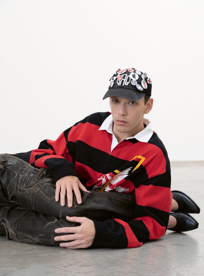 Young man with pins on cap, wearing a striped rugby shirt, is lying on top of another person against a bright white backdrop.