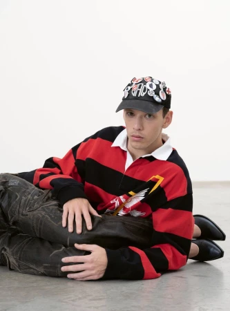 Young man with pins on cap, wearing a striped rugby shirt, is lying on top of another person against a bright white backdrop.