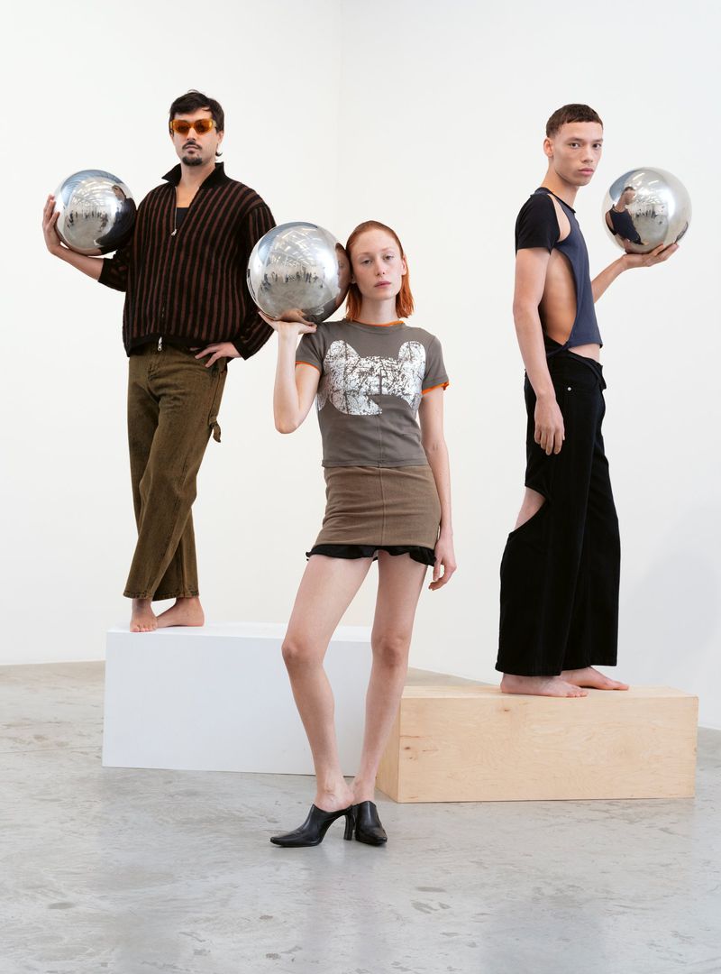 Three fashion models pose indoors on white and wood platforms, each holding a large, shiny metallic sphere in a studio setting.