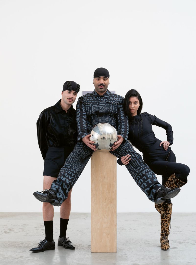 Three people in a photo studio, one man seated on a wood pillar holding a disco ball, flanked by a man and a woman in dark clothing.