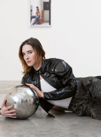Woman in a black jacket poses with a metallic sphere in an indoor setting, artfully arranged against a white wall with framed art.