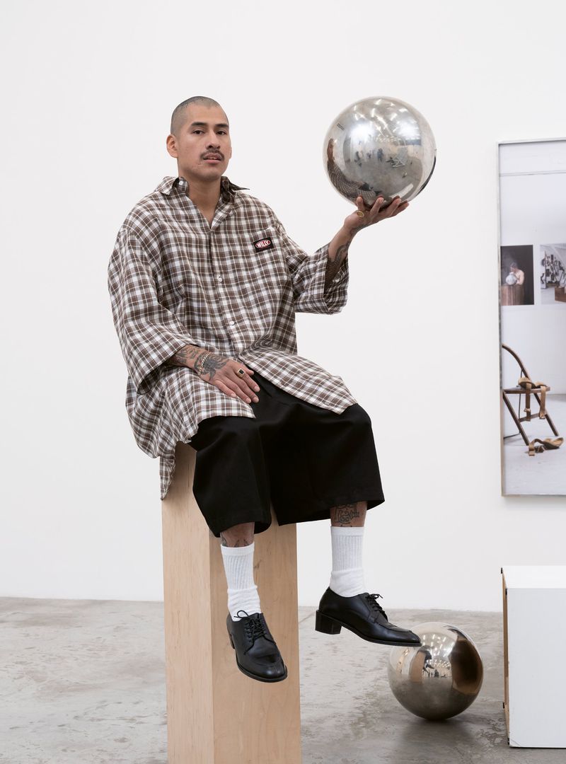 Man in oversized plaid shirt seated, holding disco ball. Wearing black shorts, white socks, and black leather shoes. Modern studio setting.
