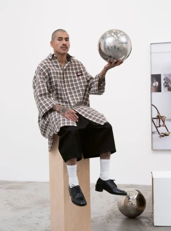 Man in oversized plaid shirt seated, holding disco ball.  Wearing black shorts, white socks, and black leather shoes.  Modern studio setting.