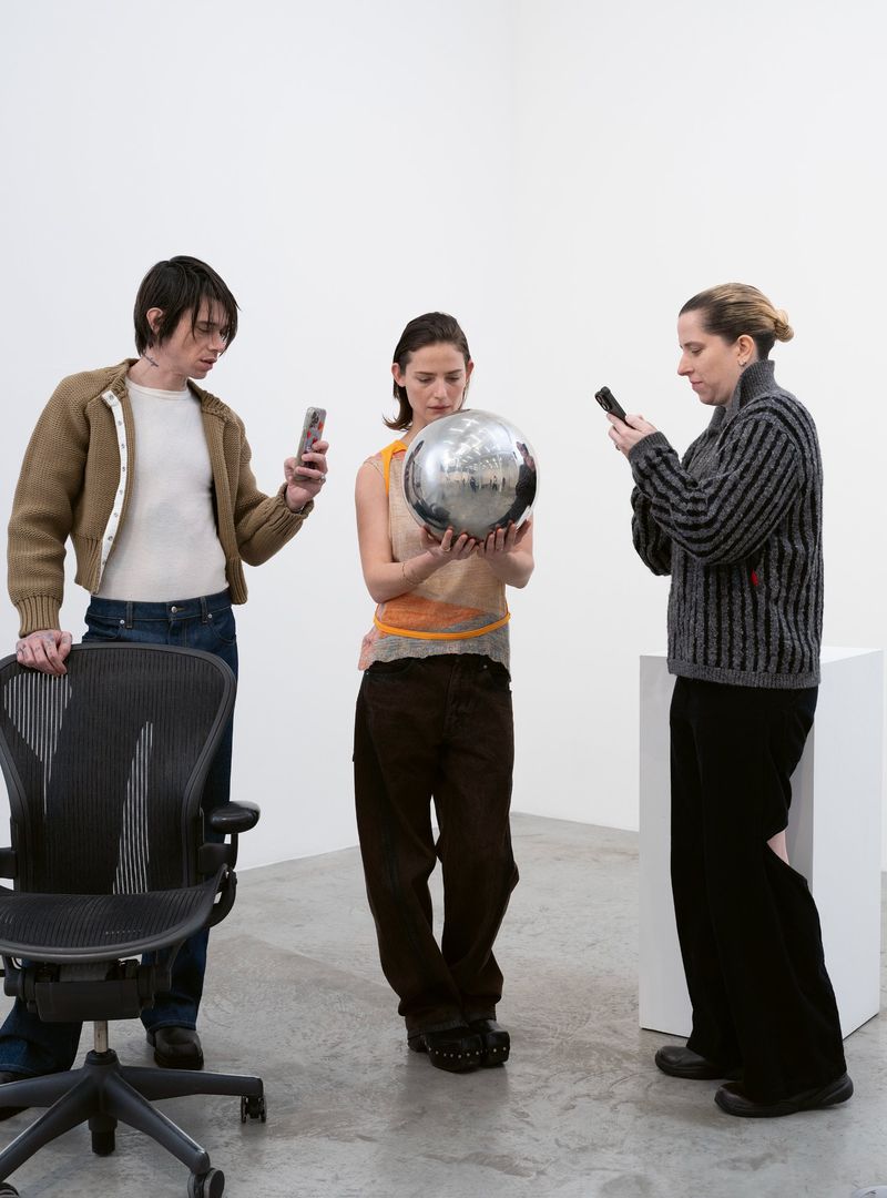 Three people in a minimalist white studio. One person holds a mirrored ball, while the other two use mobile phones, next to a chair and pedestal.