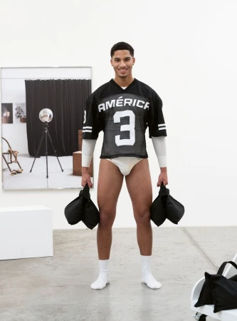 A smiling man wearing a black athletic jersey holding weighted bags in a white studio setting.