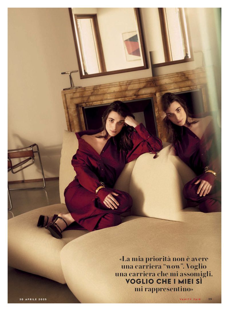 Two women in red outfits sit on a cream couch in an interior space with neutral walls, mirrors, and a chair in the background. 