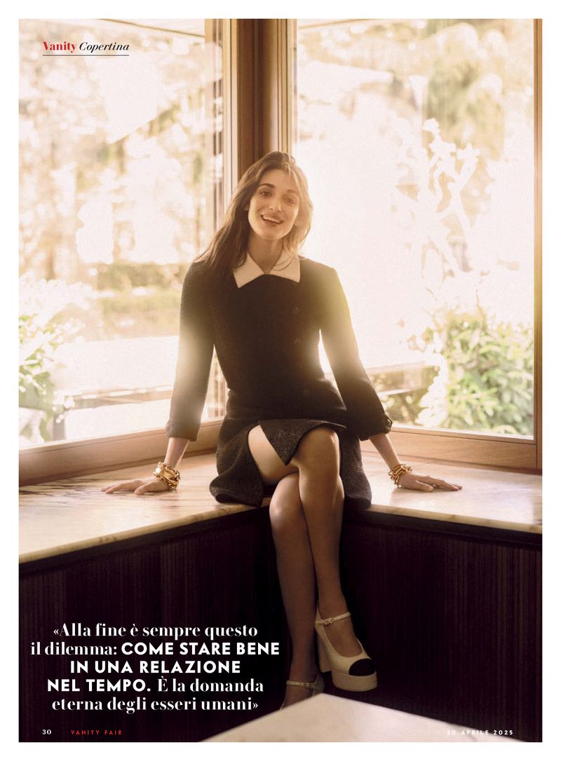 A woman in a black dress with a white collar poses seated on a window ledge in soft lighting, wearing jewelry and light-colored heels.