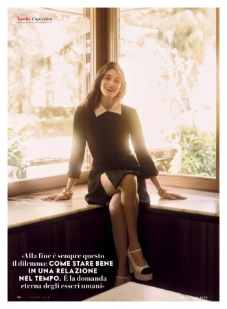 A woman in a black dress with a white collar poses seated on a window ledge in soft lighting, wearing jewelry and light-colored heels.