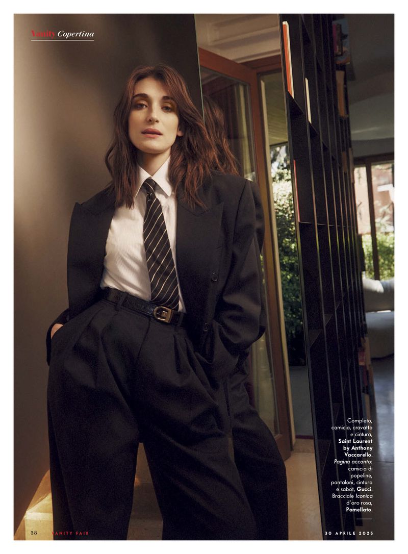 Fashion editorial shot of a brunette woman in a black suit and striped tie, standing indoors near a door and open shelving unit with neutral lighting.