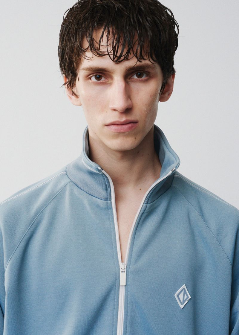 Close-up of a young adult male wearing a light blue zip-up jacket, facing the camera against a plain white backdrop in a photo studio.
