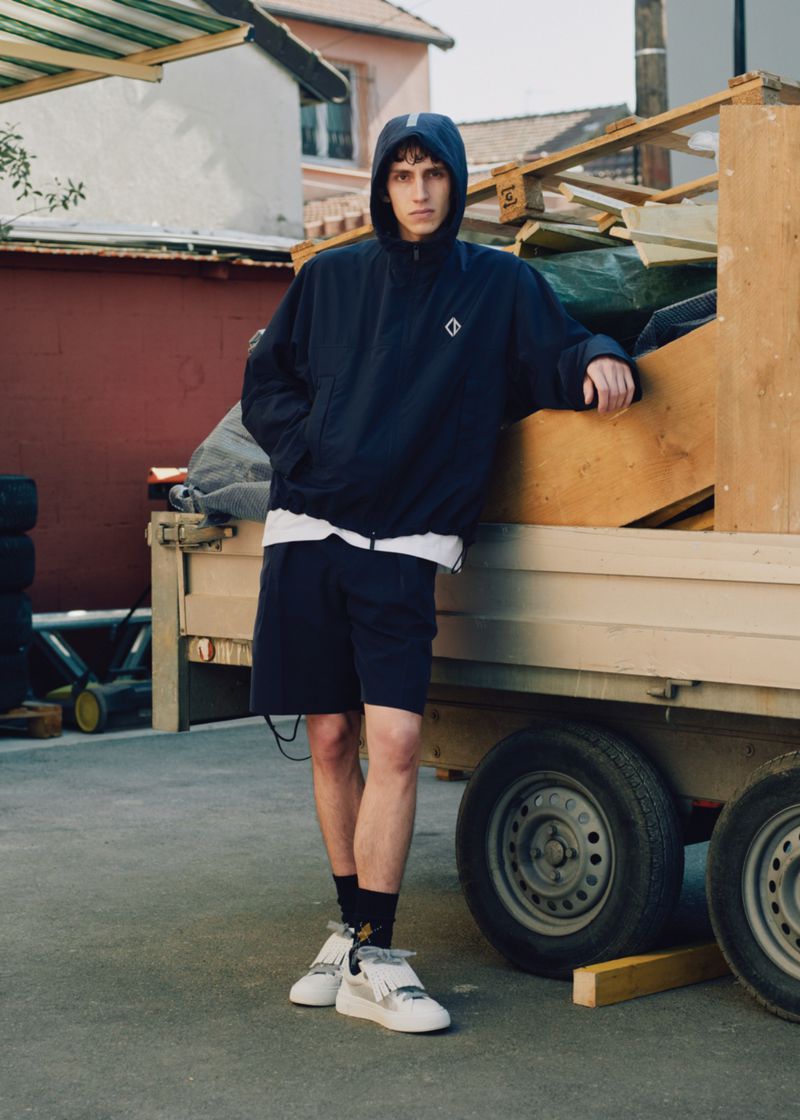 Male model in hooded jacket poses against trailer in urban setting, showcasing fashion apparel and casual outdoor style on an overcast day.