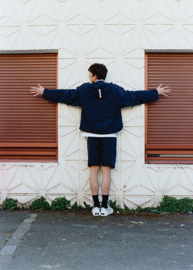 An adult poses with arms outstretched in front of a building with orange, closed window shutters in a relaxed outdoor setting.