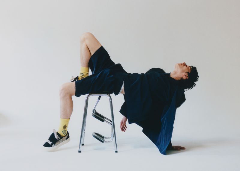 Stylish adult in relaxed pose, reclining on a chrome stool in a photo studio wearing dark casual clothes, patterned socks, and sneakers.