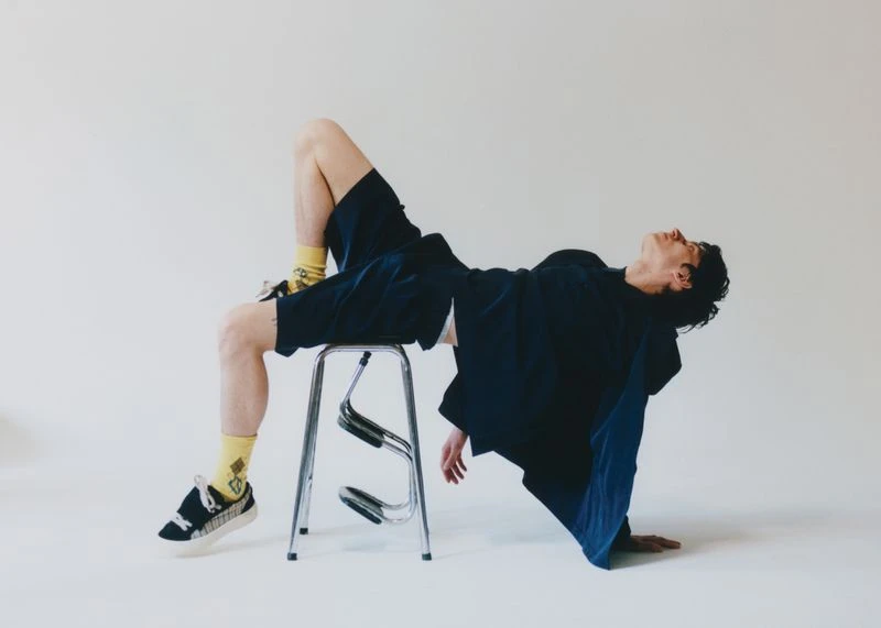 Stylish adult in relaxed pose, reclining on a chrome stool in a photo studio wearing dark casual clothes, patterned socks, and sneakers.