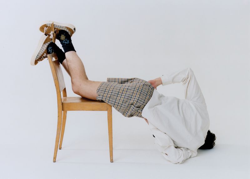 Man in plaid shorts and tan shoes balancing on chair in a bright photo studio. Artistic image with playful fashion and unique pose.