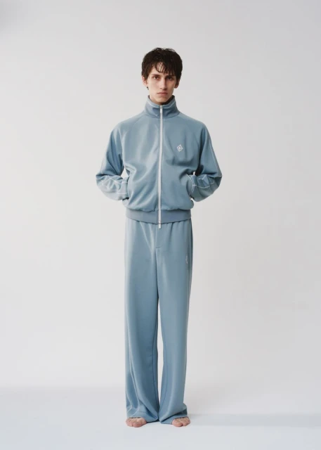 A young adult man stands in a photo studio wearing a blue zip-up tracksuit with wide-legged pants against a white background in a portrait.