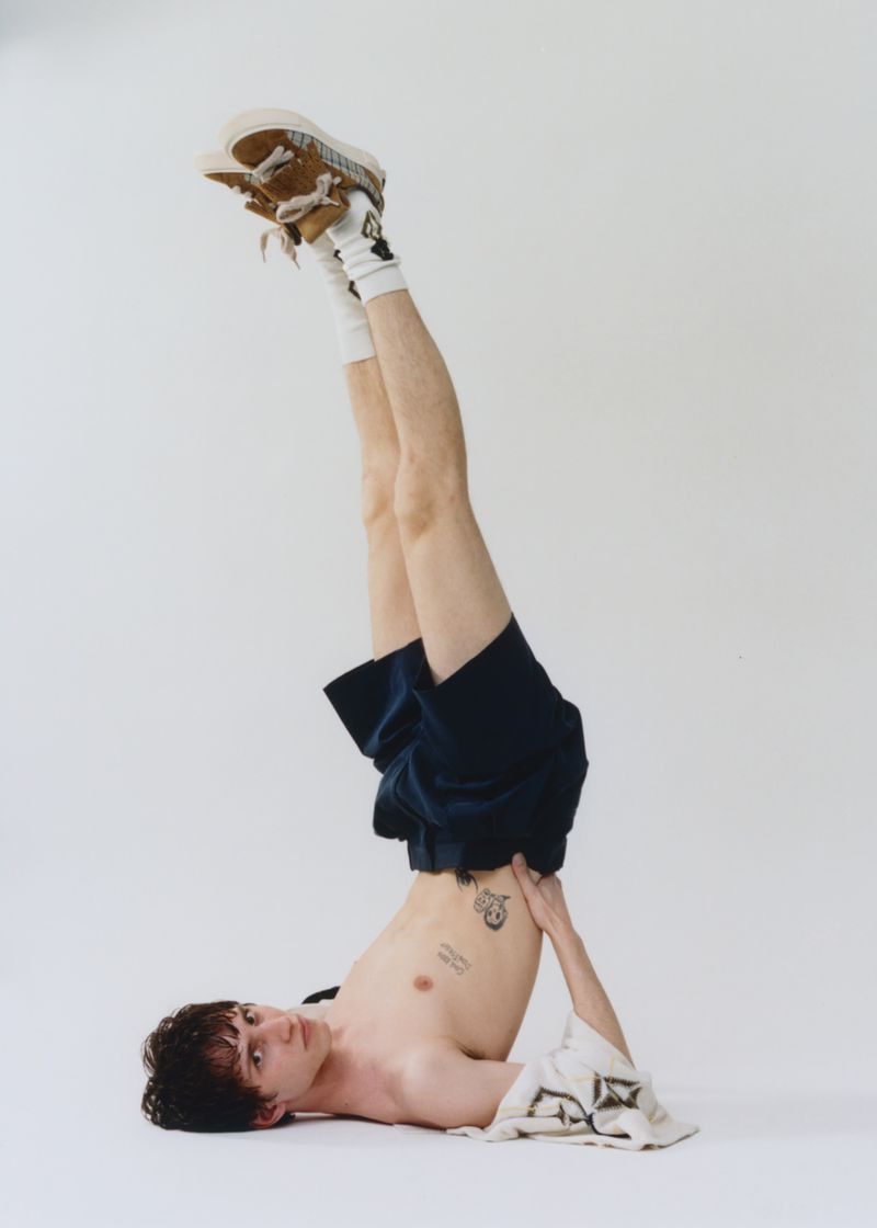 Adult male in an inverted yoga pose, legs in the air wearing shorts, socks, and shoes, with upper torso exposed in a neutral studio.