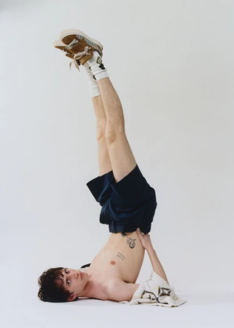 Adult male in an inverted yoga pose, legs in the air wearing shorts, socks, and shoes, with upper torso exposed in a neutral studio.