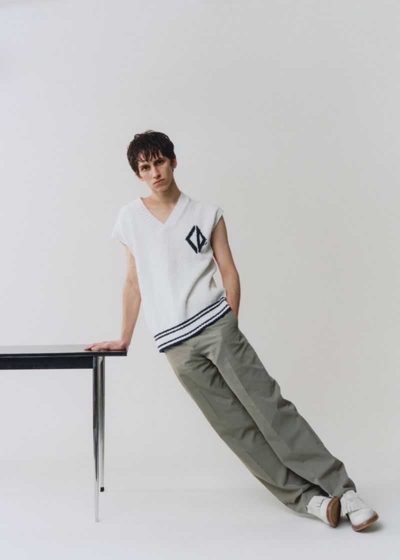 Young man in sleeveless knit top and dress pants leans on a black table in a fashion pose against a neutral studio backdrop.