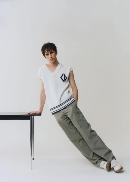 Young man in sleeveless knit top and dress pants leans on a black table in a fashion pose against a neutral studio backdrop.
