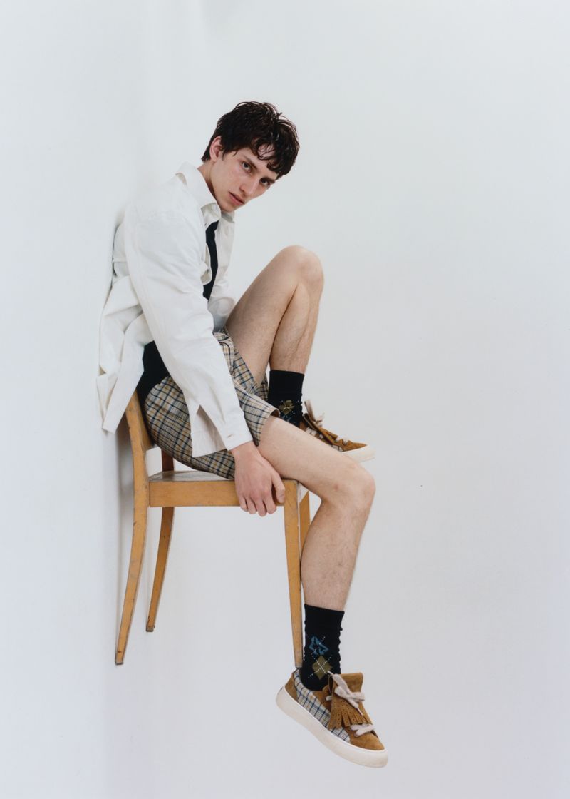 Young male model in a relaxed pose, seated on a chair, wearing a white shirt, plaid shorts, and sneakers against a white background.
