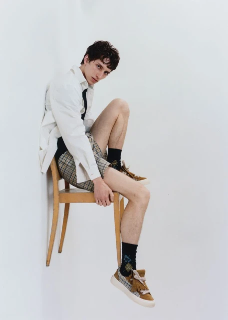 Young male model in a relaxed pose, seated on a chair, wearing a white shirt, plaid shorts, and sneakers against a white background.