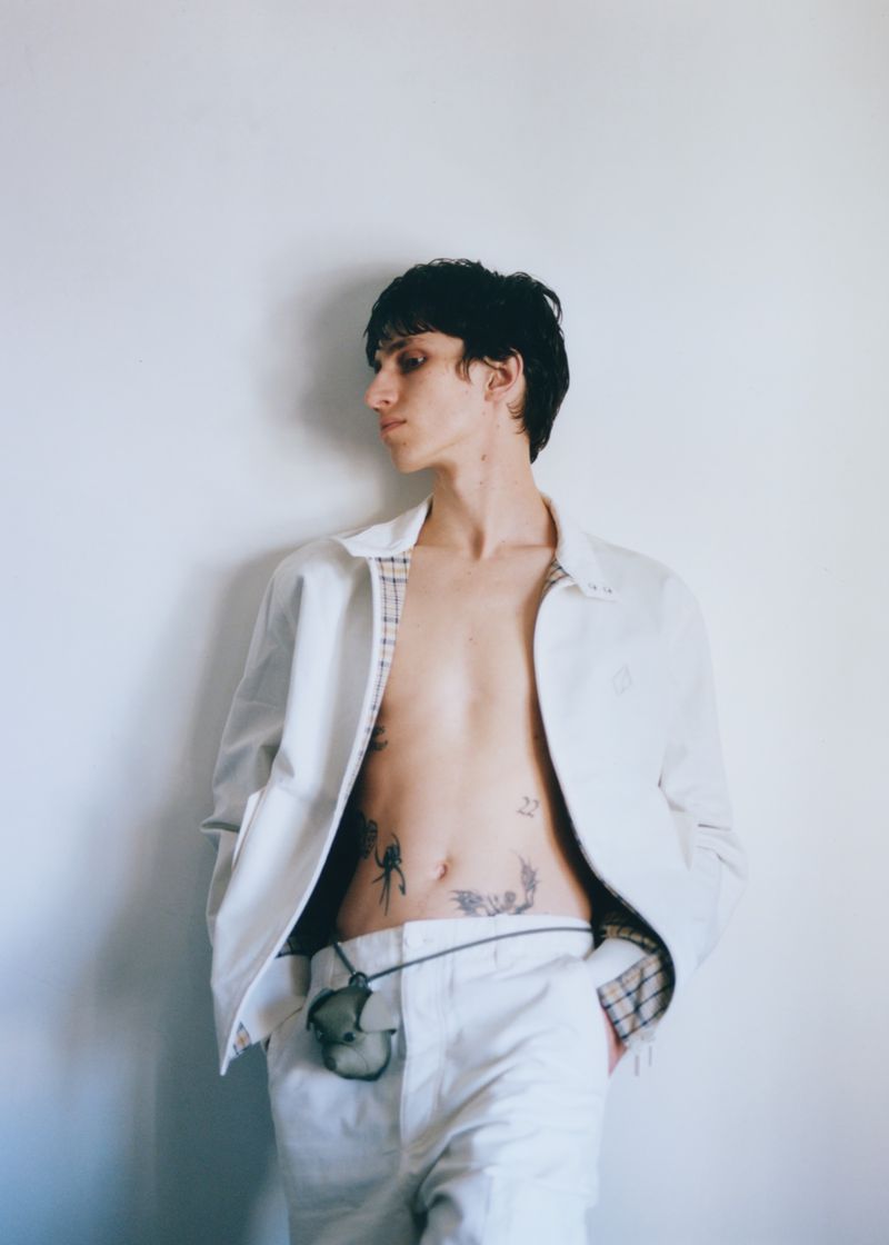 A stylish young adult stands in a white jacket, light pants, and a pendant necklace against a pale background, capturing a fashion-forward moment.